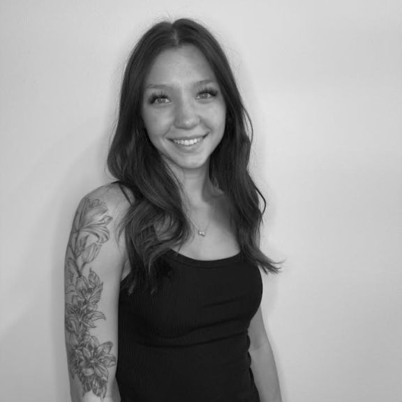 Woman with long hair wearing a sleeveless top and a floral arm tattoo, standing against a plain wall and smiling. Black and white photo.