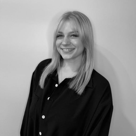 A young woman with straight, light hair wearing a dark button-up shirt stands smiling in front of a plain light-colored wall. The image is in black and white.
