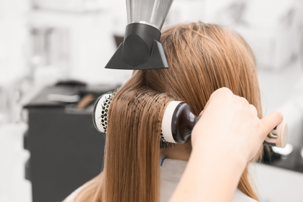 Person’s hair is being styled with a round brush and blow dryer at a salon, focusing on smoothing and straightening the strands.