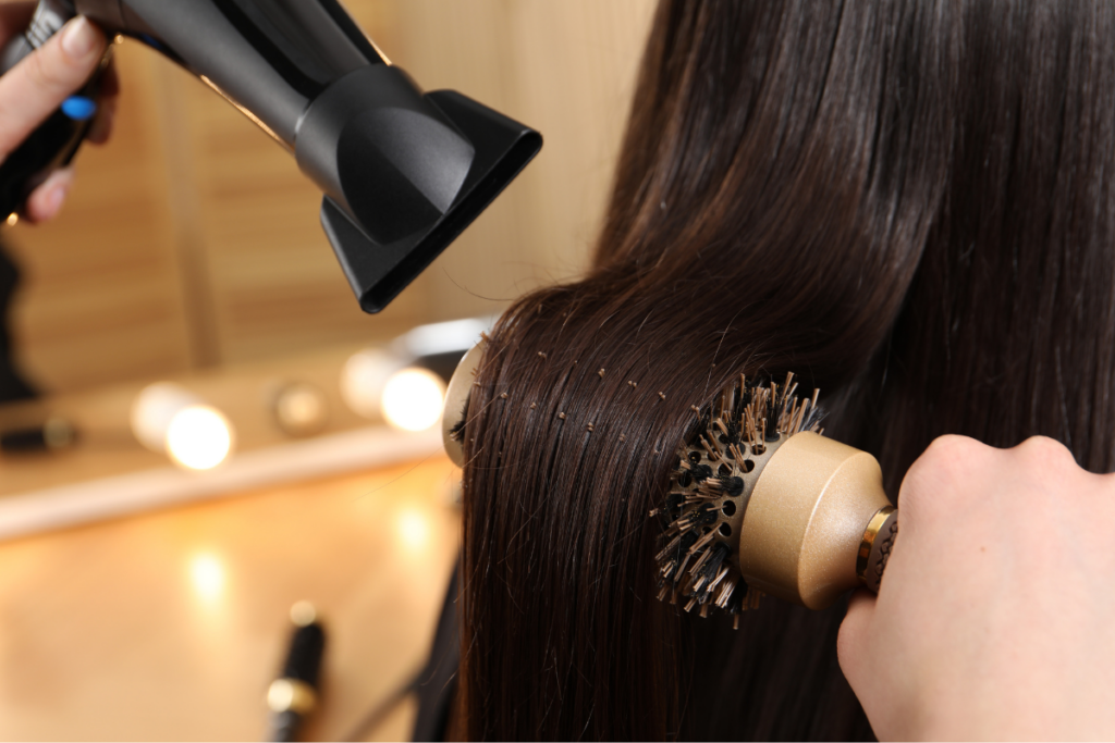 Close-up of a hair dryer and round brush positioned against long dark hair with blurred lights in the background.