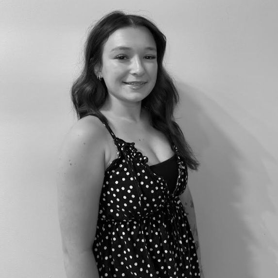 A person with long hair wearing a polka dot sleeveless top stands against a plain wall, facing the camera and smiling slightly.