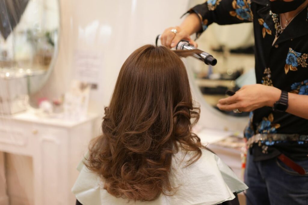 A hairstylist uses a curling iron to style the hair of a seated person in a salon.