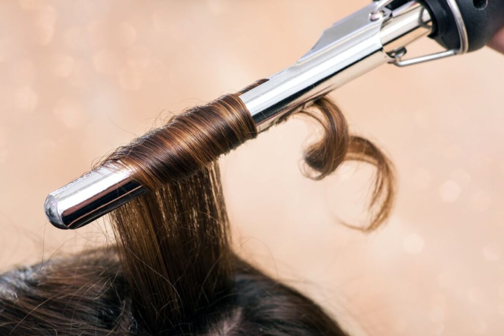 A close-up of brown hair being curled around a silver curling iron against a soft, blurred background.