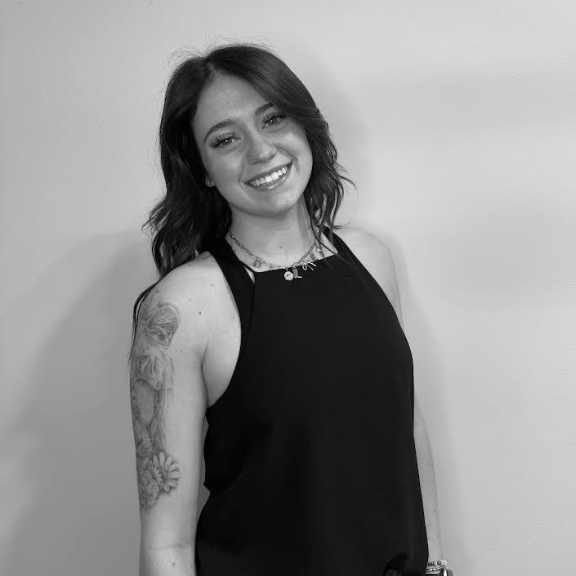 Woman with long dark hair, wearing a sleeveless black top, smiles at the camera. She has a floral tattoo on her left upper arm and stands against a plain light-colored wall.