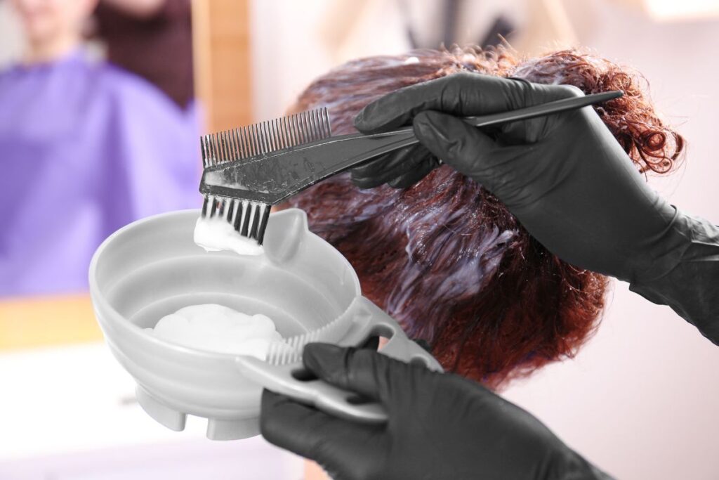 Person wearing black gloves applies hair dye with a brush to someone’s hair, using a plastic bowl containing white cream.