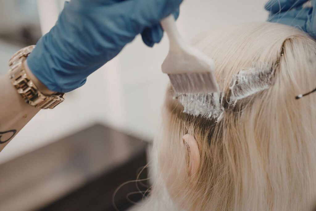 A person wearing blue gloves applies hair dye or bleach to the roots of blonde hair with a brush.