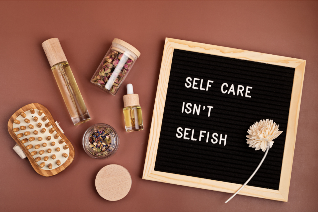A wooden massage brush, glass bottles, dried flowers, a flower stem, and a letter board displaying the words “SELF CARE ISN’T SELFISH" on a pink background.