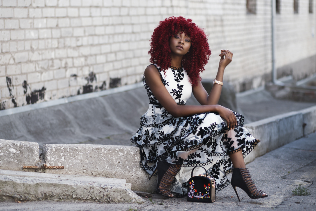 Person with bright curly red hair sitting on a concrete ledge outdoors, wearing a black-and-white patterned dress and high-heeled sandals.
