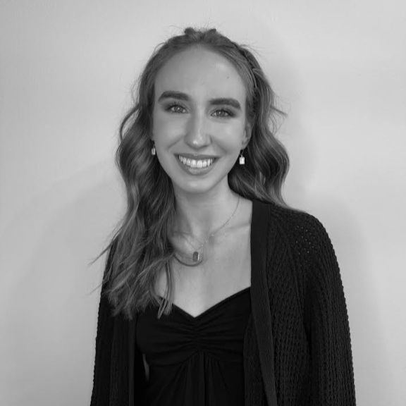 A young woman with long wavy hair, wearing a dark top and cardigan, smiles at the camera in front of a plain light-colored wall. The image is in black and white.