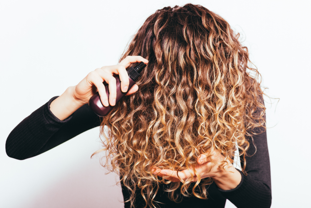 Person holding a spray bottle near long wavy hair while supporting the ends of the hair with one hand against a light background.