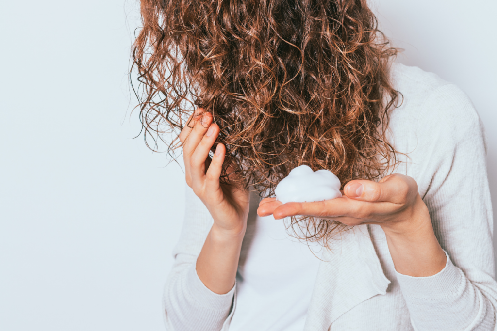 A person with curly hair holds a dollop of foam or mousse in their hand, preparing to apply it to their hair—perfect for following an extensions guide to achieve fuller, bouncier curls.
