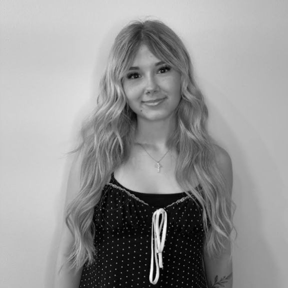 A young woman with long, wavy hair stands against a plain wall, wearing a sleeveless, polka-dot top and a necklace. The image is in black and white.
