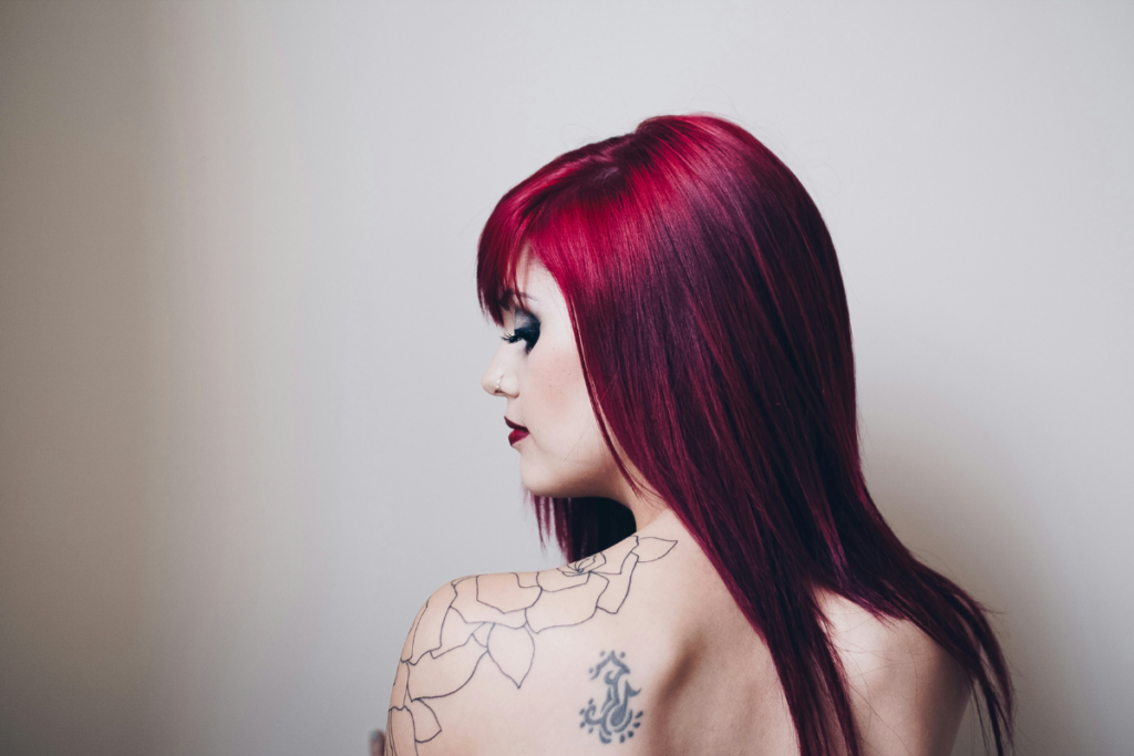 Side profile of a person with straight burgundy-red hair, wearing dark eye makeup and showing shoulder tattoos against a light background.
