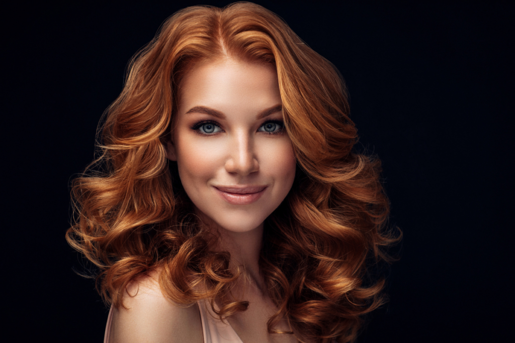 Portrait of a smiling person with shoulder-length copper-colored wavy hair against a dark background.