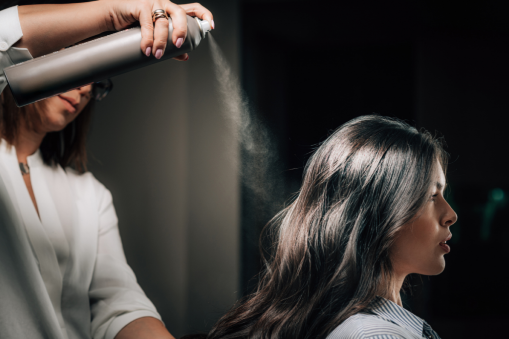 Woman holding an aerosol can that is releasing a fine mist toward another woman with long dark hair.