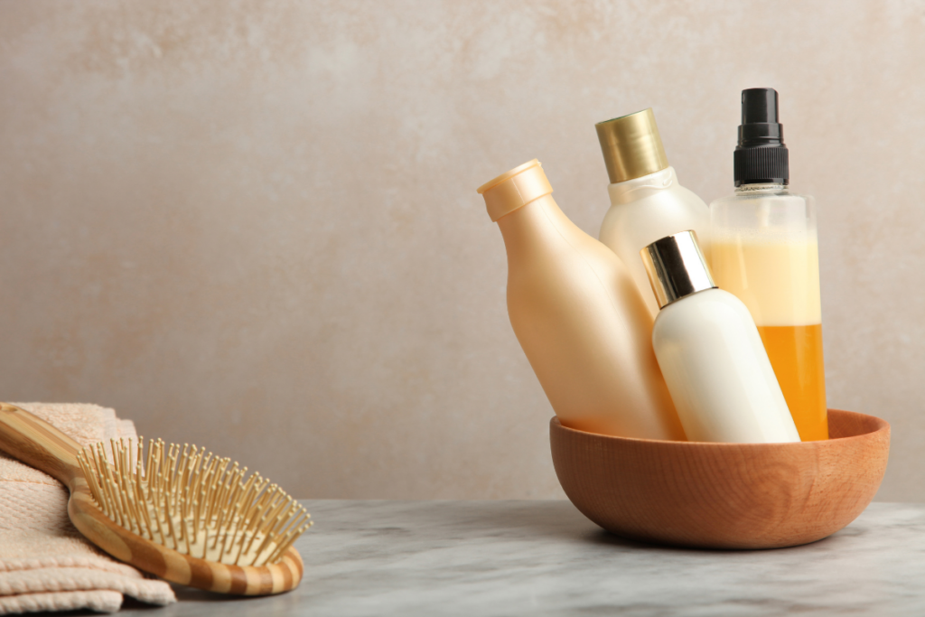 A wooden bowl with assorted shampoo and conditioner bottles next to a wooden hairbrush and folded towels on a marble surface.