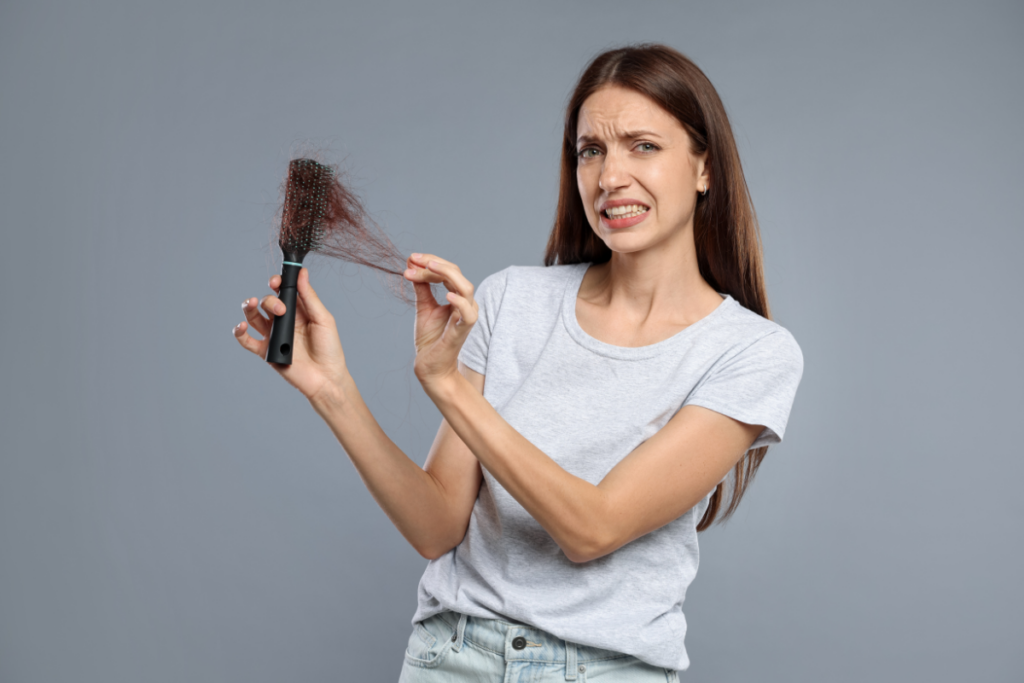 A woman in a gray t-shirt looks concerned while holding a hairbrush full of tangled hair against a plain gray background.