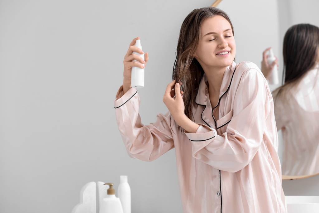 Woman in striped pajamas sprays a product onto her wet hair in a bathroom, standing near a counter with bottles and a mirror, following an extensions guide for optimal hair care.