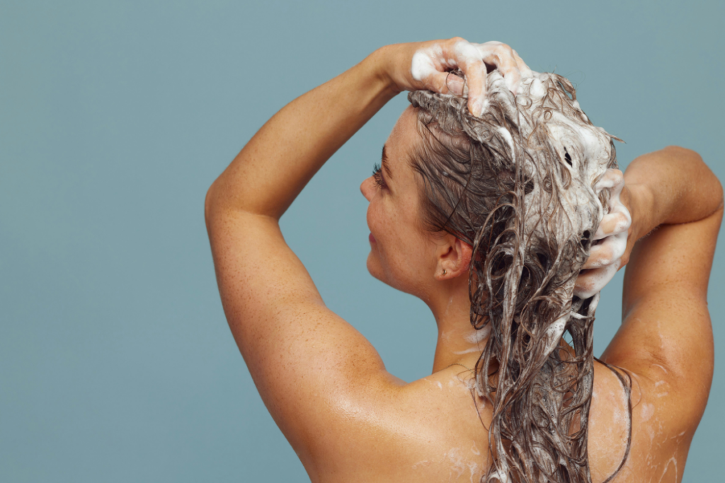 Woman with wet hair covered in shampoo suds while both hands massage the scalp against a blue background.