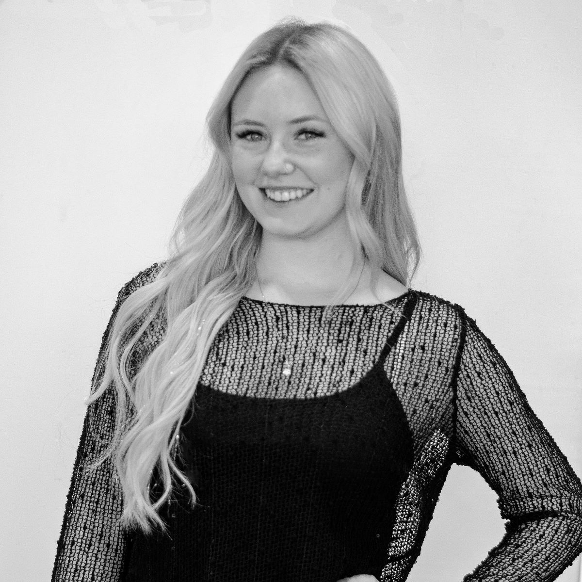 A woman with long blonde hair, wearing a black mesh top, stands and smiles against a plain background.