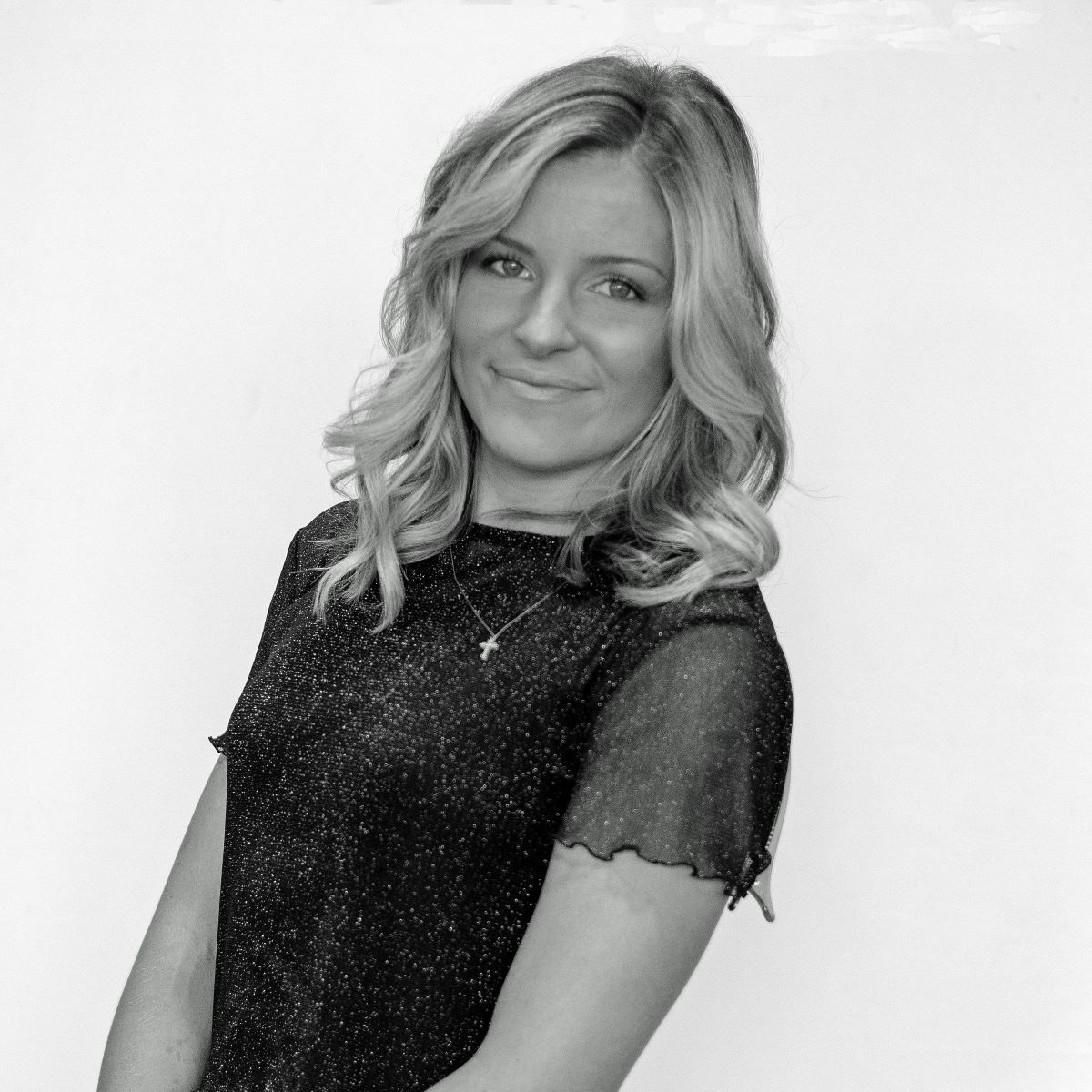 Black and white photo of a person with shoulder-length wavy hair wearing a sparkly short-sleeve top and necklace, standing against a plain background.