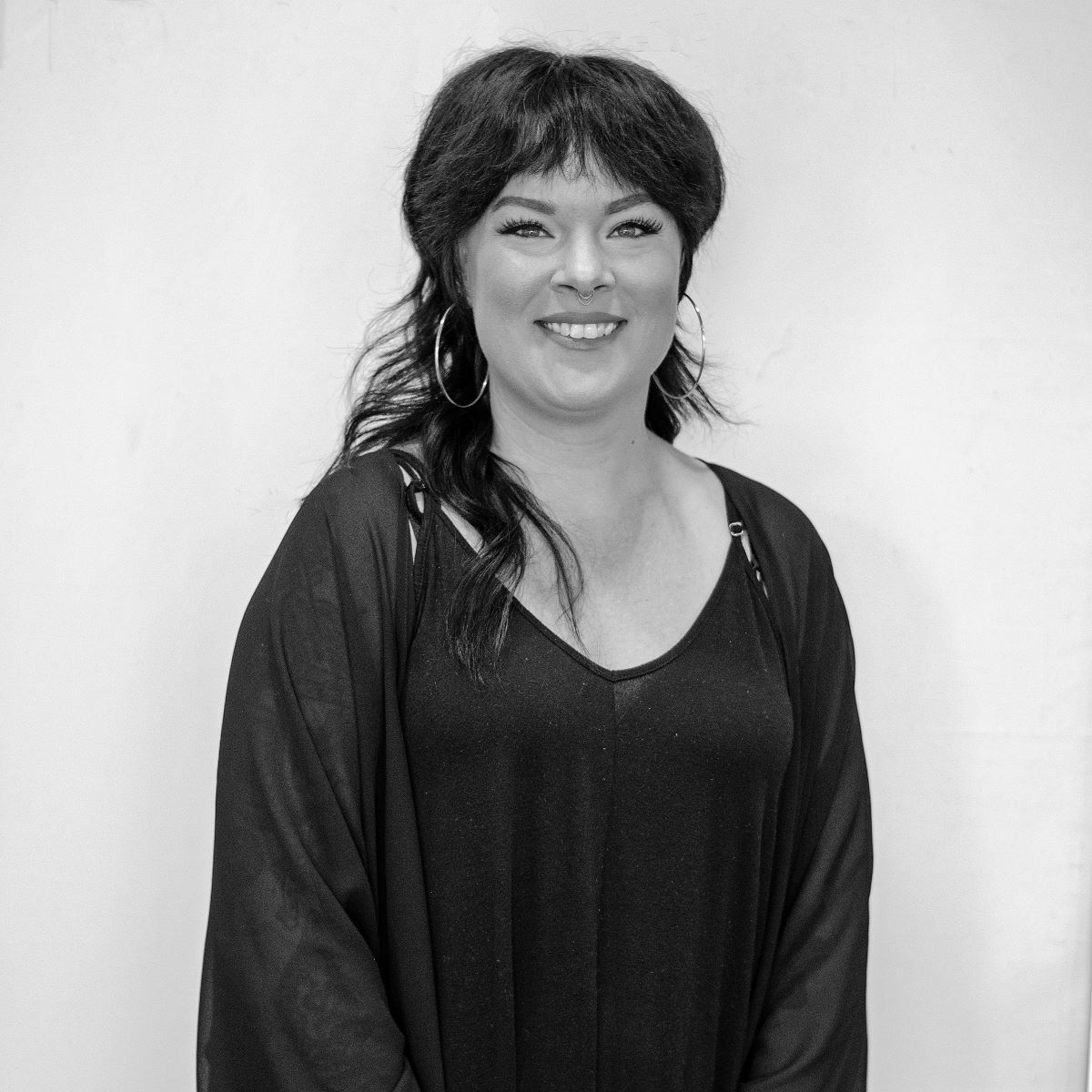 A woman with dark hair and hoop earrings smiles while standing against a plain white background. She is wearing a black top and a black cardigan.