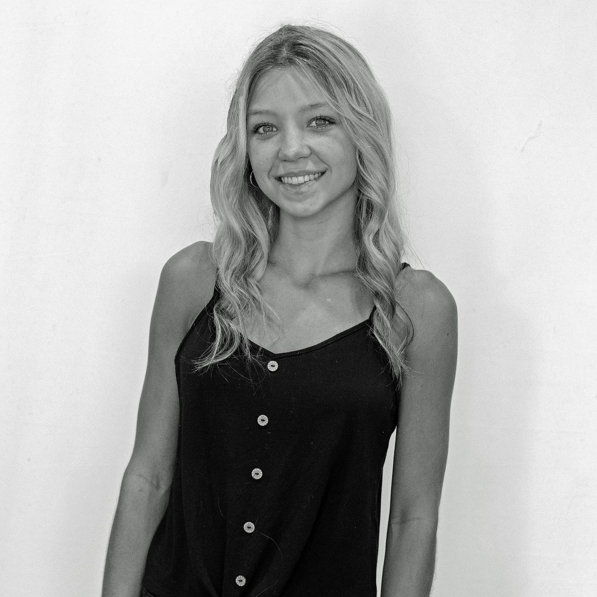 A woman with long, wavy hair wearing a sleeveless button-up top smiles at the camera against a plain background.