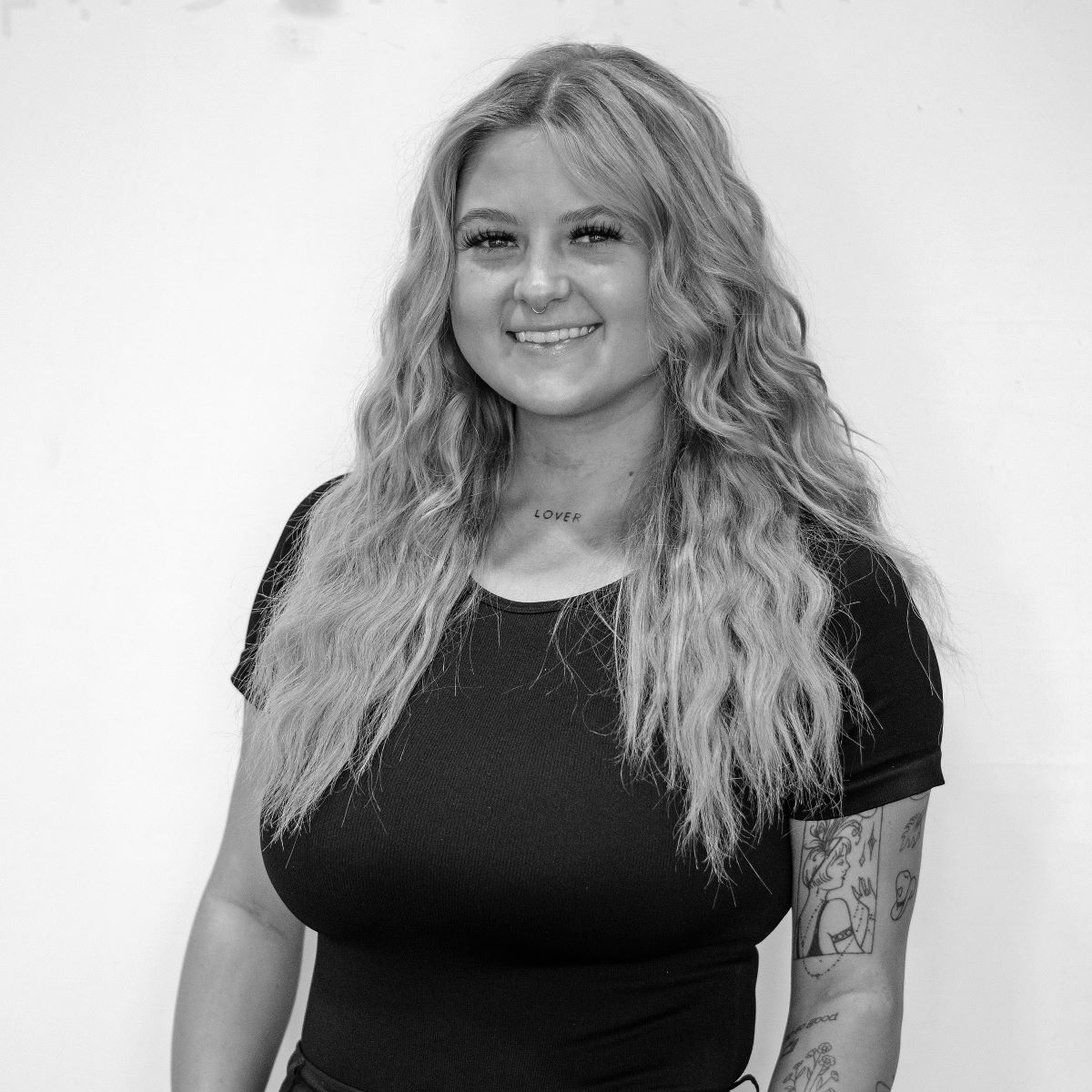 A person with long, wavy hair wearing a black t-shirt smiles for the camera. Tattoos are visible on their left arm and neck.