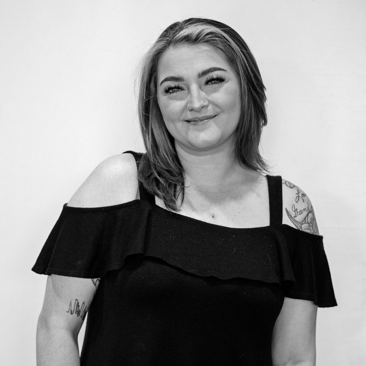 A woman with shoulder-length hair and tattoos on her left arm is posing against a plain background. She is wearing an off-the-shoulder black top and smiling.