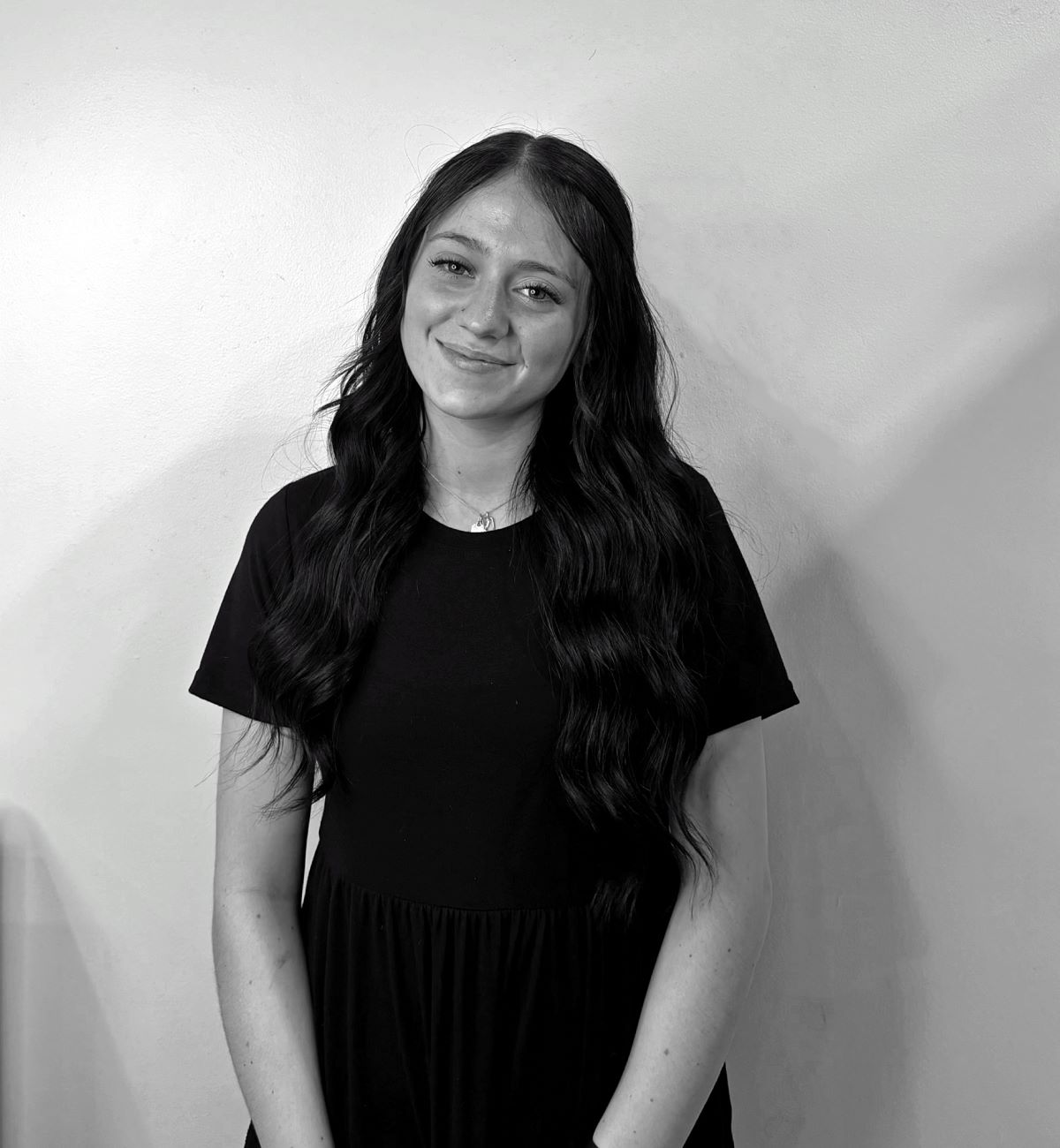 A woman with long wavy hair wears a black dress and stands against a plain wall, smiling. The photo is in black and white.