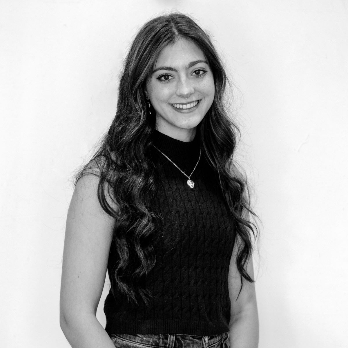 Black and white photo of a smiling person with long, wavy hair, wearing a sleeveless, textured top and a necklace, standing against a plain background.