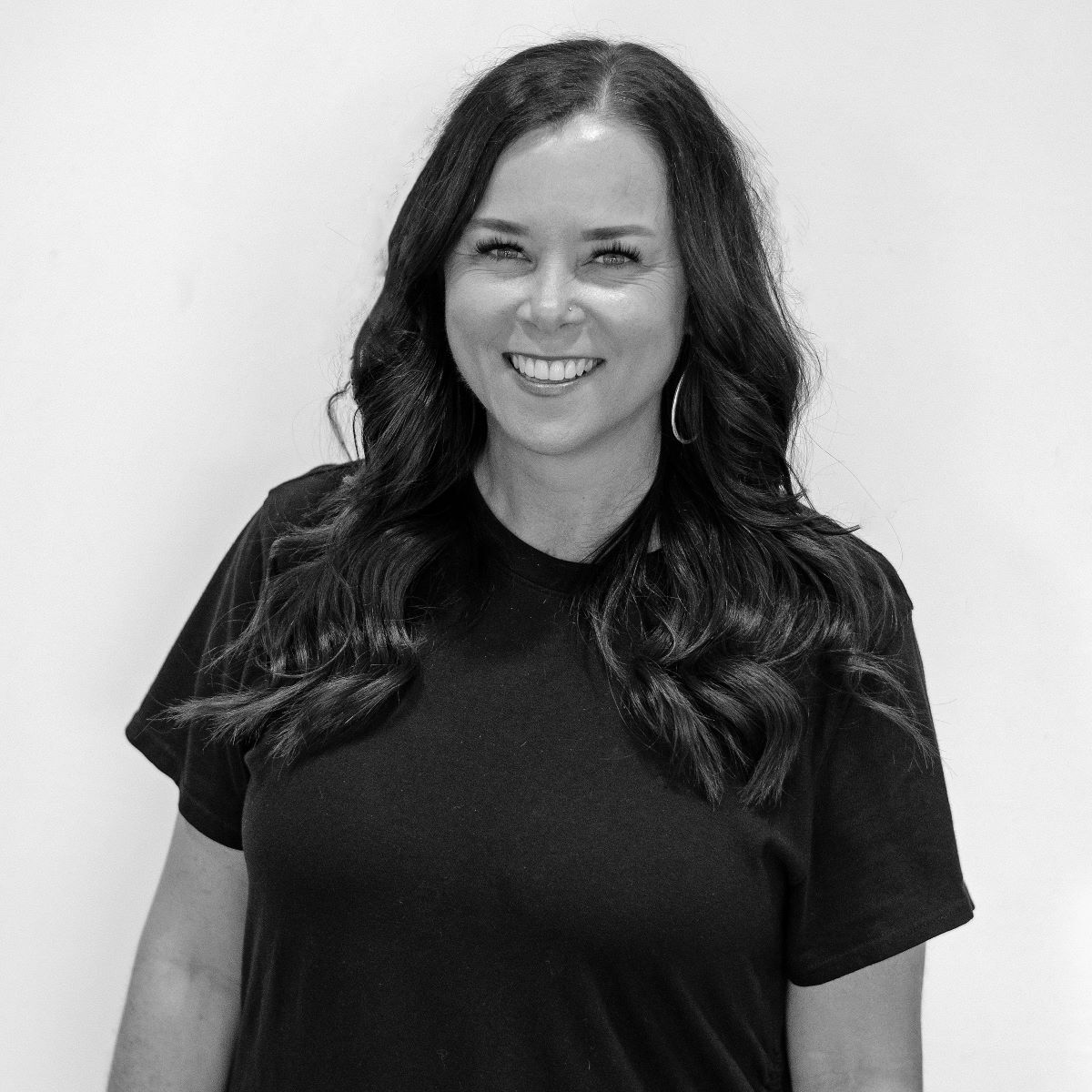 A woman with long dark hair and a black t-shirt smiles while facing the camera against a plain background.