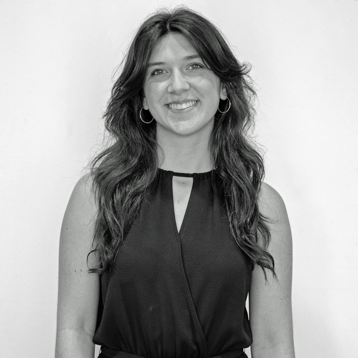 Black-and-white photo of a smiling person with long wavy hair wearing a sleeveless, dark-colored top. The background is plain and light-colored.