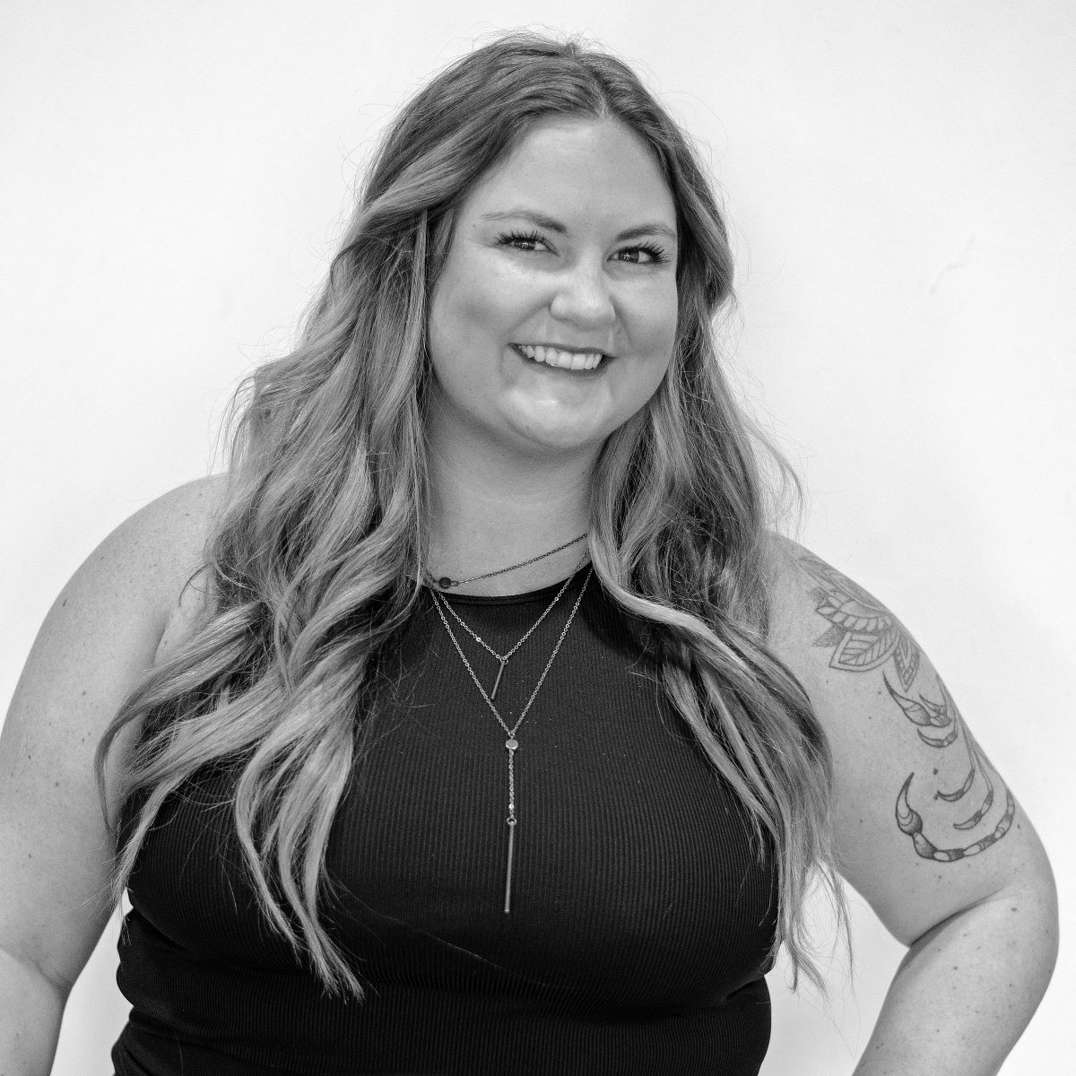 Black and white portrait of a smiling person with long wavy hair, wearing a sleeveless top and layered necklaces, and showing tattoos on their right arm.