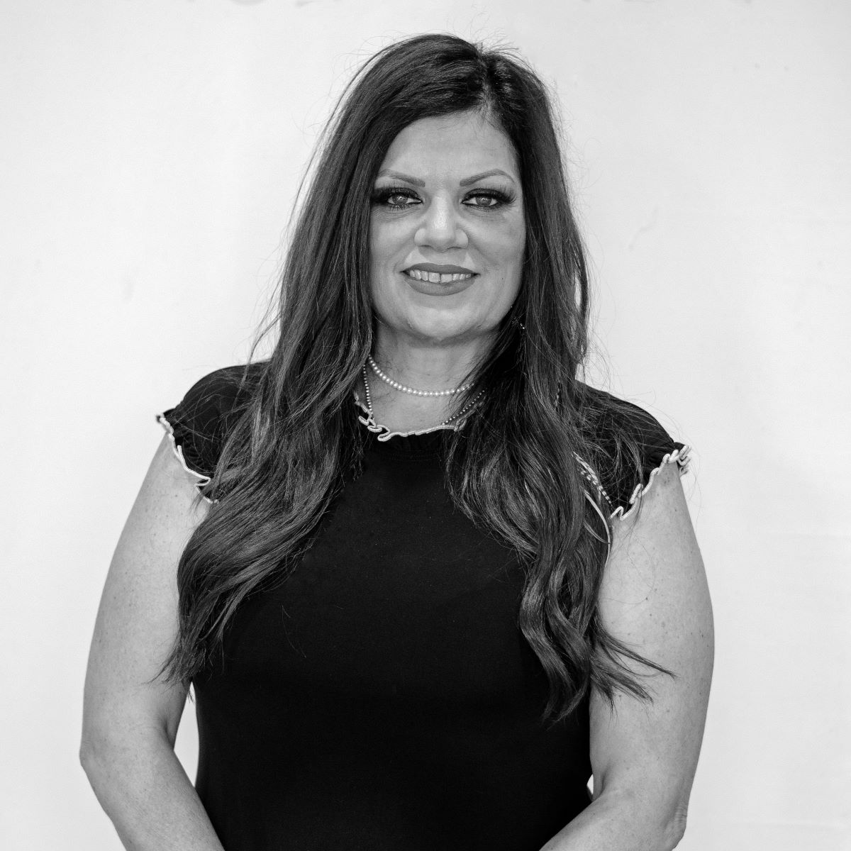 A woman with long dark hair wearing a short-sleeved, black top stands against a plain background, smiling at the camera.