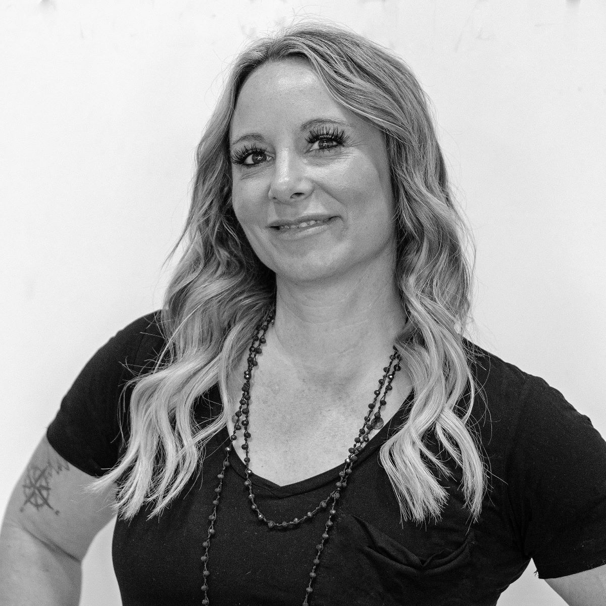 Black and white image of a woman with long wavy hair, wearing a black top and layered necklaces, smiling slightly at the camera. A tattoo of a pentagram is visible on her left arm.