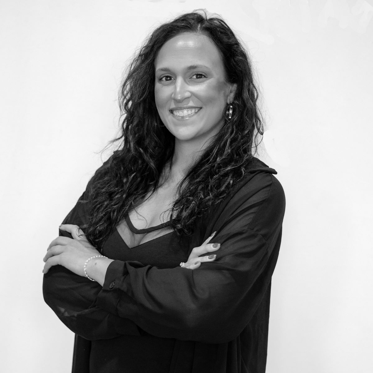 Black and white photo of a woman with long curly hair, smiling and standing with her arms crossed. She is wearing a dark top and light jewelry.